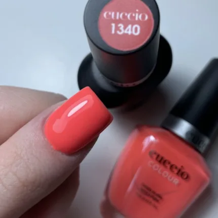 CUCCIO 1340 lakier tradycyjny 13 ml – swatch koralowego lakieru na paznokciu obok butelki z czarną nakrętką